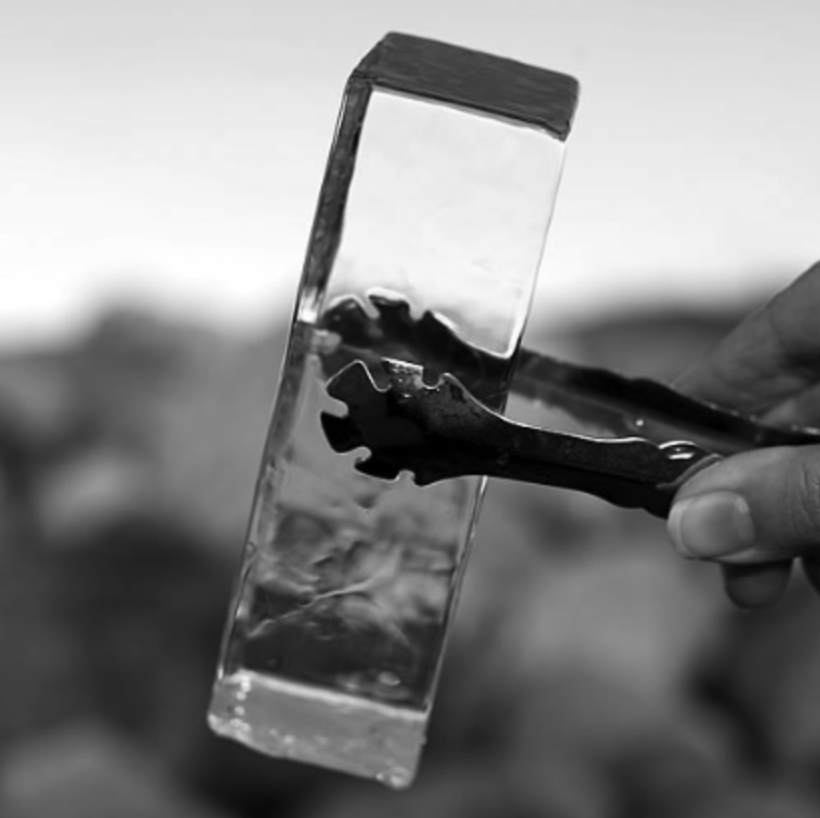 Black-and-white close-up of a rectangular clear ice cube held by metal tongs, with water droplets glistening on its surface against a blurred background.
