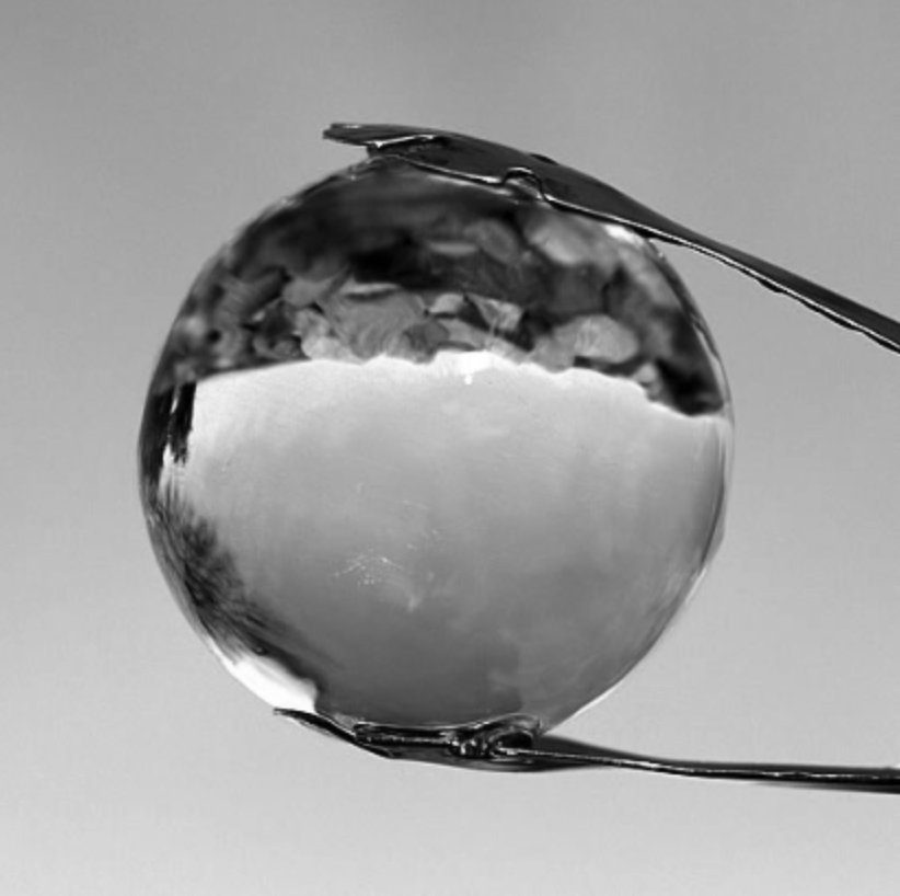 Black-and-white close-up of a spherical clear ice cube held by metal tongs, with water droplets glistening on its surface against a blurred background.